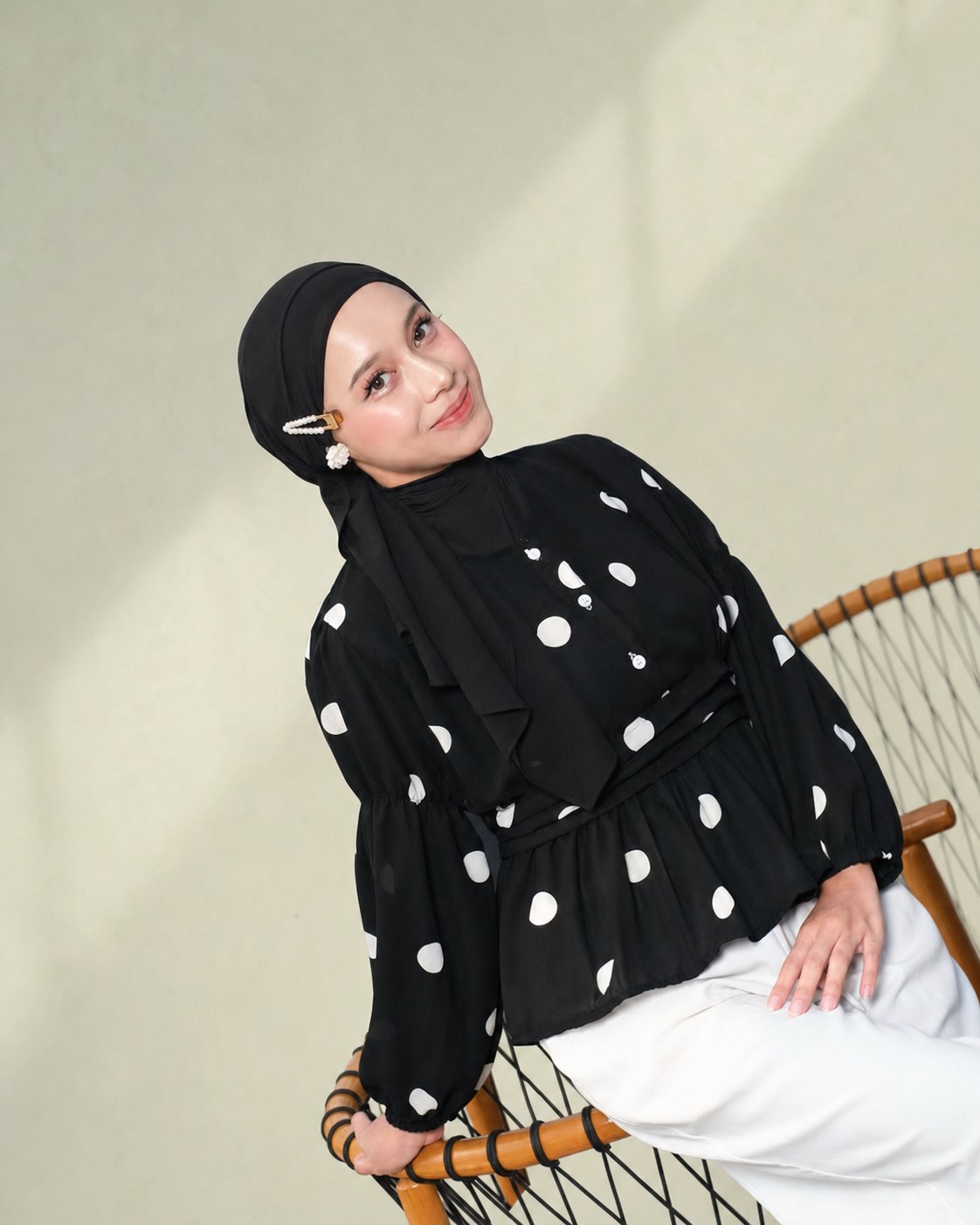 Model berhijab hitam mengenakan atasan hitam polkadot dan celana putih