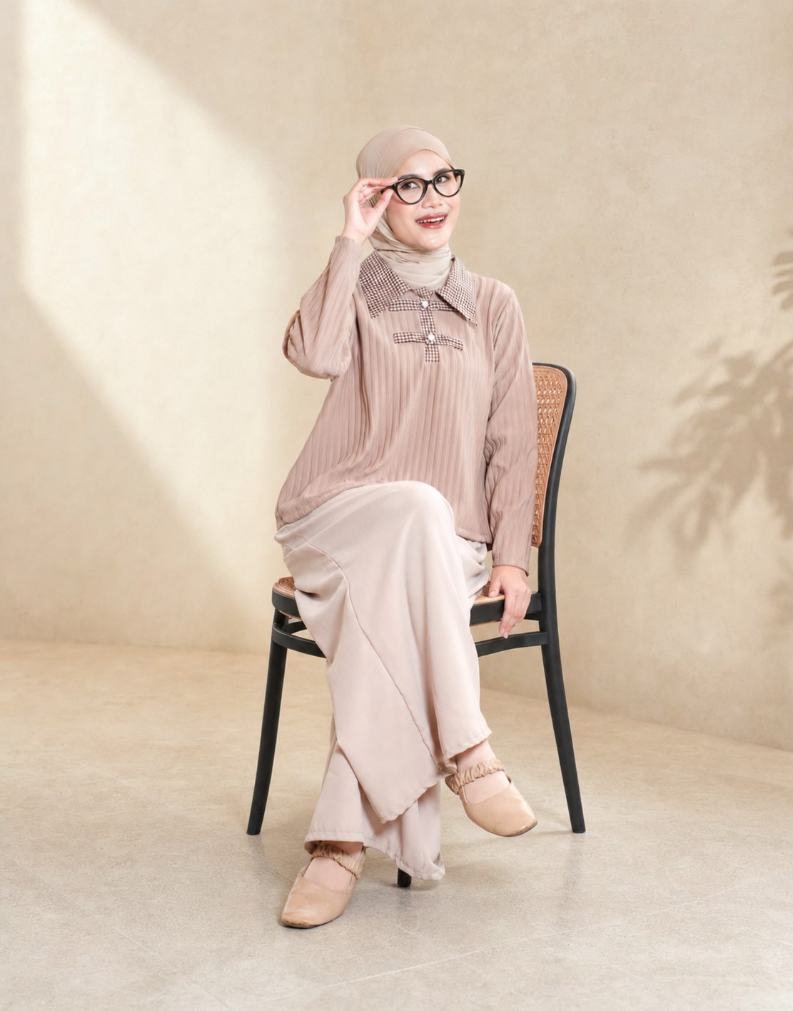 Model berhijab beige dan berkacamata mengenakan busana taupe sambil duduk
